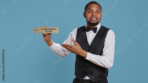 Hotel concierge using a wall indicator to guide clients to the conference room, presenting various amenities at the five star luxury resort. Bellboy staff holds a sign for directions. Camera B. photo