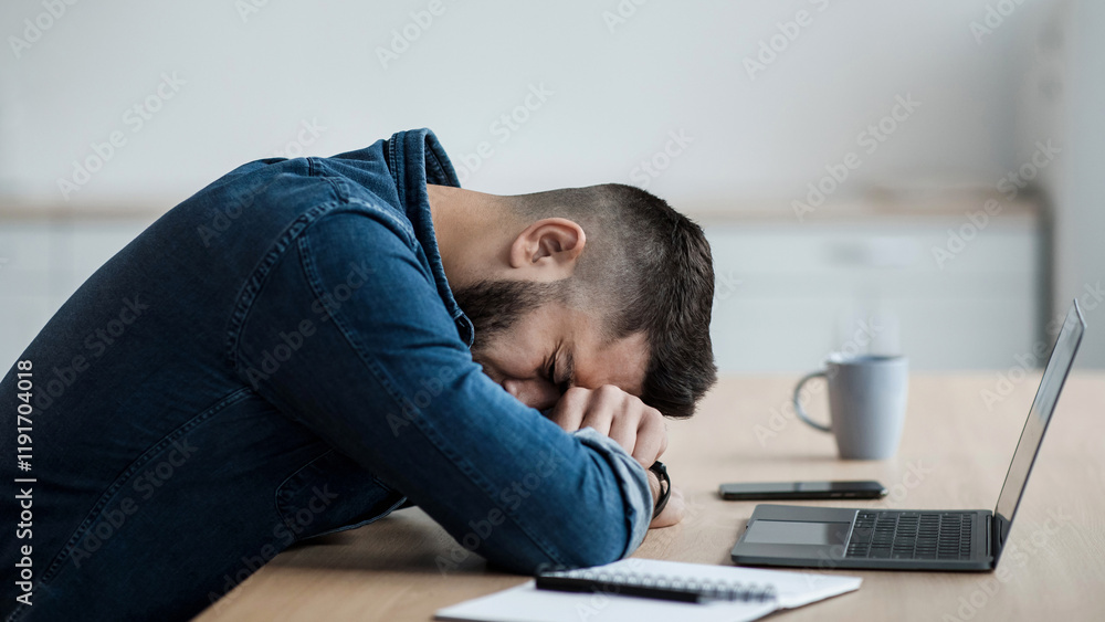 Fototapeta premium Problems at work, mistake, fatigue, dismissal, stress and overwork. Sad depressed tired millennial man lies on desk, suffers from tension at table with laptop, in kitchen interior, copy space