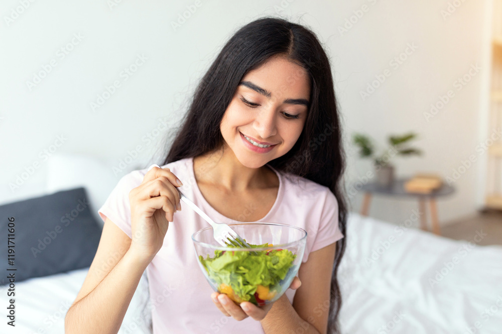 Healthy vegan diet concept. Joyful Indian lady sitting on bed at home with bowl of fresh vegetable salad, eating tasty lunch. Pretty Eastern woman enjoying her vegan meal, promoting clean living Healthy vegan diet concept. Joyful Indian lady sitting on bed at home with bowl of fresh vegetable salad, eating tasty lunch. Pretty Eastern woman enjoying her vegan meal, promoting clean living