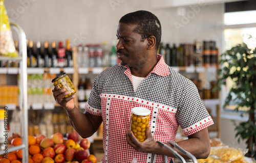 African man looking at canned pickled olives in store. Market visitor choose consider jar with marinated olives. Buyer pick up set of groceries, consider available options, makes choice before buying photo