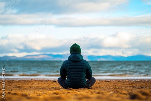 A person meditates peacefully on a sandy beach, facing the tranquil ocean and distant mountains, embodying tranquility and mindfulness in a stunning natural landscape. photo