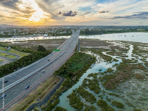 Aerial view of motorway traffic over a coastal wetland at sunset. Cars travel on the highway, while boats are moored in the marina. NORTHWESTERN MOTORWAY, AUCKLAND, NEW ZEALAND photo