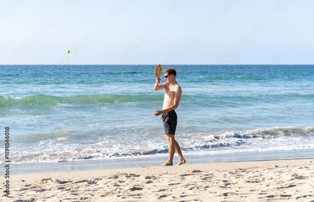 A blonde young man in swimwear and sunglasses enjoying a game of paddle ball on the beach under the sunny sky.