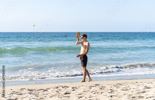Obraz na płótnie A blonde young man in swimwear and sunglasses enjoying a game of paddle ball on the beach under the sunny sky. w salonie
