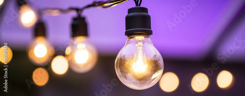 Glowing light bulb with string lights bokeh against purple twilight creating nostalgic illuminated ambiance for outdoor event photo