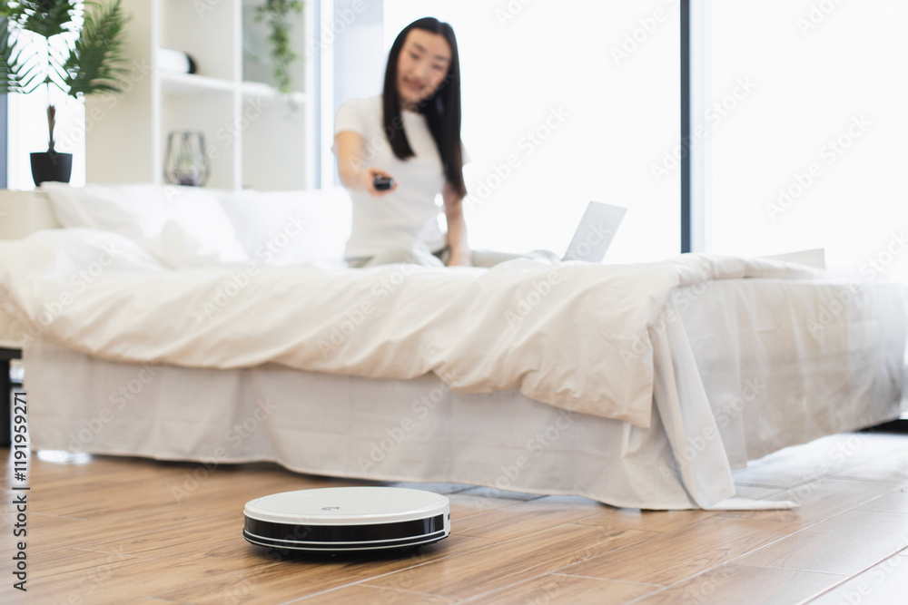 Young Asian woman in bedroom controlling robot vacuum cleaner. Concept of automated cleaning, smart home technology, and modern lifestyle. Young Asian woman in bedroom controlling robot vacuum cleaner. Concept of automated cleaning, smart home technology, and modern lifestyle.