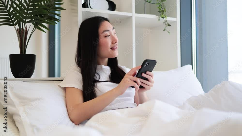 Young Asian woman relaxing on bed using smartphone. Modern bedroom with natural light and greenery creates calming atmosphere.