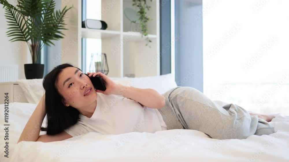 Asian woman relaxing on bed having cheerful phone conversation. She is wearing casual clothes and appears content in a bright, cozy room. Asian woman relaxing on bed having cheerful phone conversation. She is wearing casual clothes and appears content in a bright, cozy room.