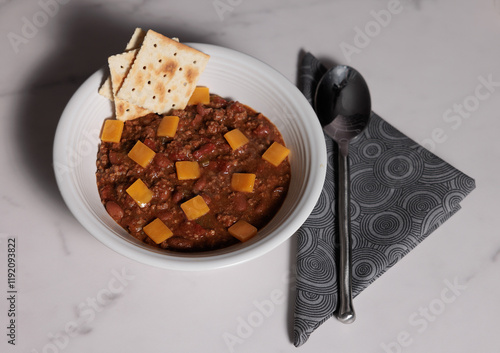 Bowl of chili with cheese and crackers next to a spoon on a napkin