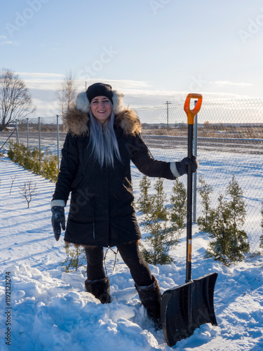 Happy woman dressed in warm winter clothes, energetically removing snow with shovel in bright winter landscape. Working diligently to clear snow-covered garden winter day outdoors in winter sunlight. 