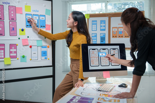 Two female web designers engaging in a lively discussion about user interface design for a mobile application, utilizing a whiteboard and computer in a collaborative office environment photo