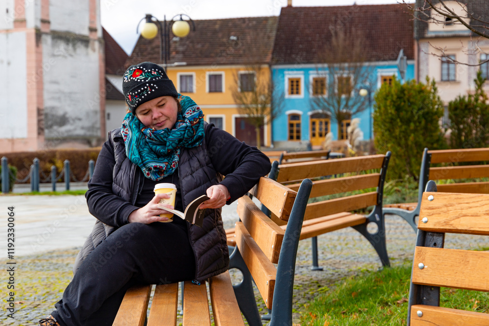 A woman sitting on a bench, reading a book, and drinking coffee.