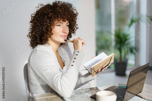 Thoughtful woman sitting with book and pen at home photo