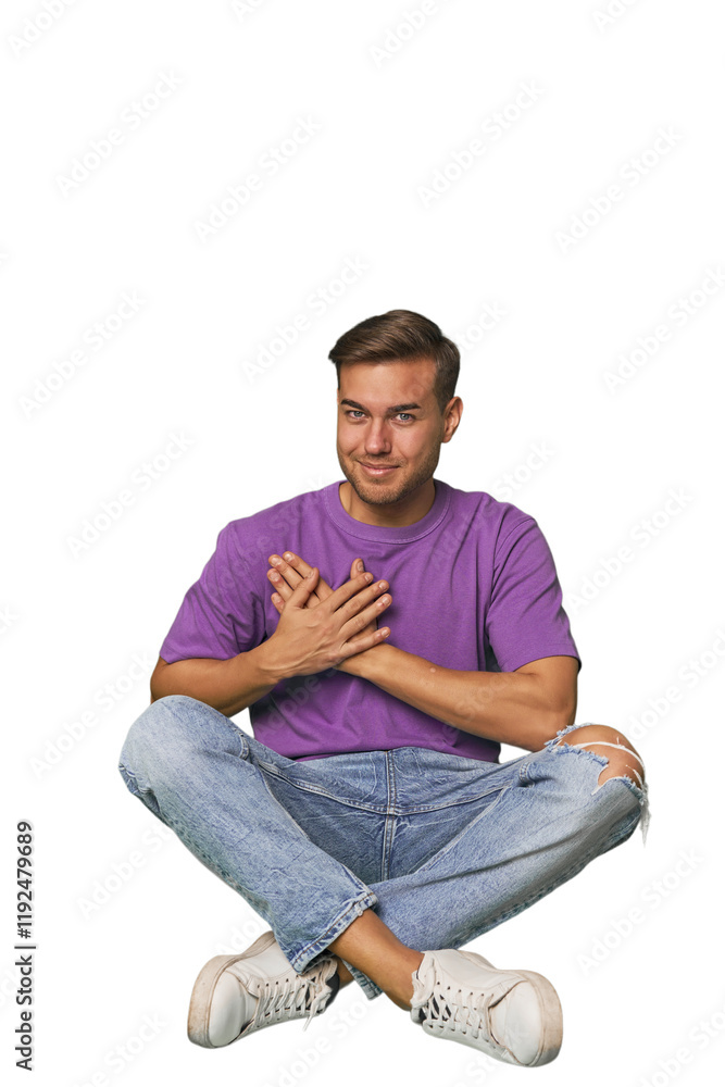 Casual man sitting on floor has friendly expression, pressing palm to chest. Love concept. Casual man sitting on floor has friendly expression, pressing palm to chest. Love concept.