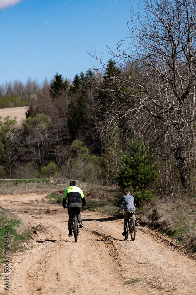 Two people bike along a sandy path. Trees and foliage surround the path. The sky is clear and blue. It's a pleasant, sunny day for a bike ride. Two people bike along a sandy path. Trees and foliage surround the path. The sky is clear and blue. It's a pleasant, sunny day for a bike ride.