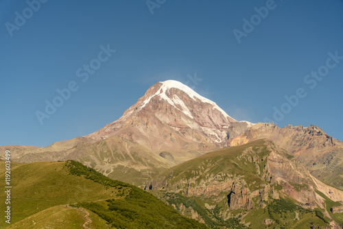 A mountain with a snow covered peak and a clear blue sky. The mountain is surrounded by green grass and trees photo