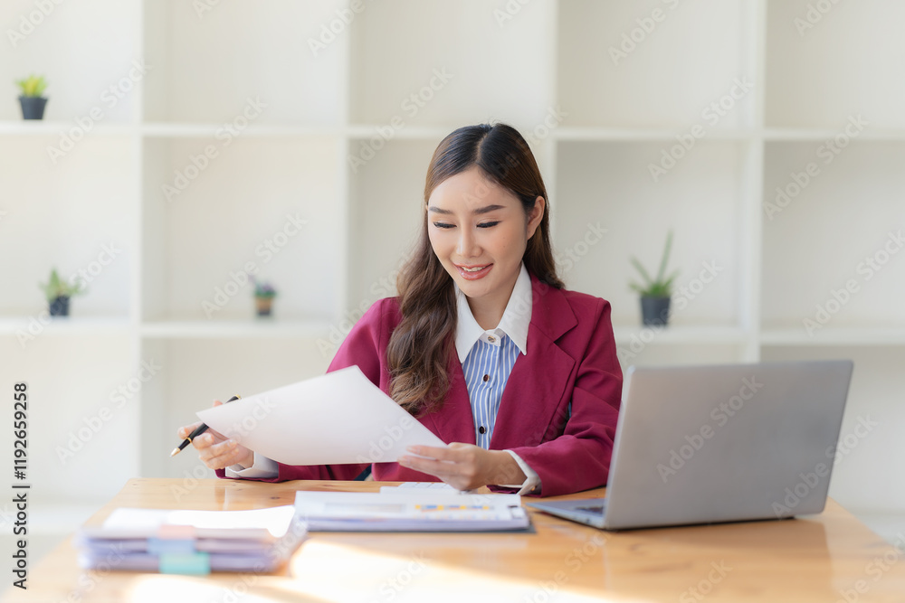 Smiling Asian accountant working with calculator and analyzing financial documents on laptop at table surrounded by documents on table Smiling Asian accountant working with calculator and analyzing financial documents on laptop at table surrounded by documents on table