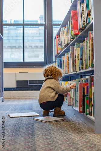 Toddler with curly hair selecting books from a shelf in a library, with a book open on the floor. Cleveland, Ohio, USA photo