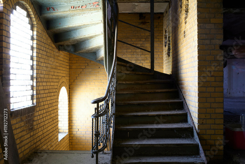 Sunlight filters through an empty building's  window onto the yellow brick wall of a stairwell with a black ornate railing and blue painted ceiling, Berlin, Germany photo