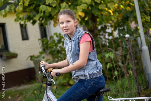 Teenage girl in a denim vest riding a bicycle on a sunny day near a house with green foliage, Netherlands photo