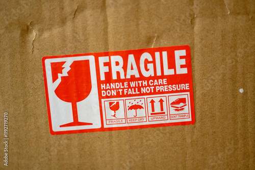 Red and white fragile label on a cardboard box, featuring icons and text for handling instructions: fragile, keep dry, upward, don't trample