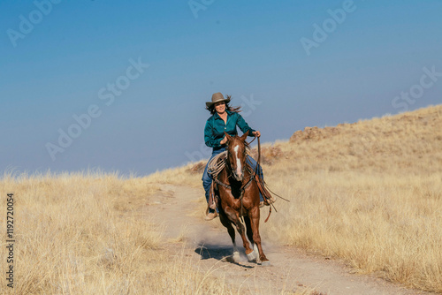 A woman wearing a cowboy hat rides a brown horse swiftly along a dirt path surrounded by dry, golden grass under a clear blue sky, Idaho, USA