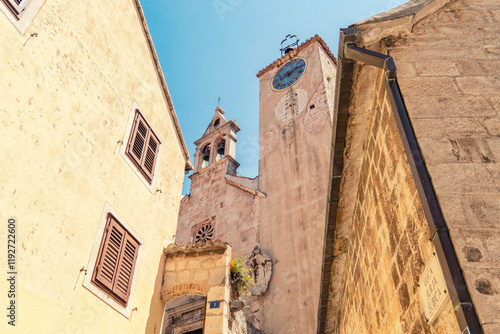 Historic buildings with shuttered windows flank narrow streets leading to an aged clock tower of Church of the Holy Spirit   in Omis under a clear blue sky, Croatia photo