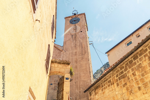 Sunlit old clock tower  of the Church of the Holy Spirit in Omis, between historic stone buildings under a clear blue sky, Croatia
