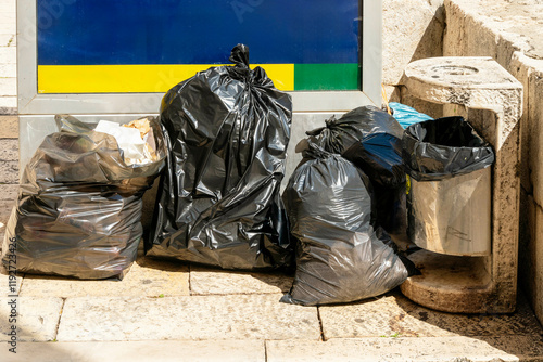 Black garbage bags piled beside a stone waste container on a sunny day with an informational sign in the background, Split, Croatia photo