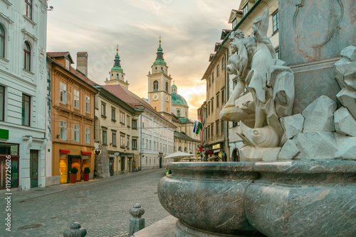 Sunset illuminates an old European street with a foreground focus on a stone fountain and historic St. Nicholas Church in the background, Ljubljana, Slovenia photo