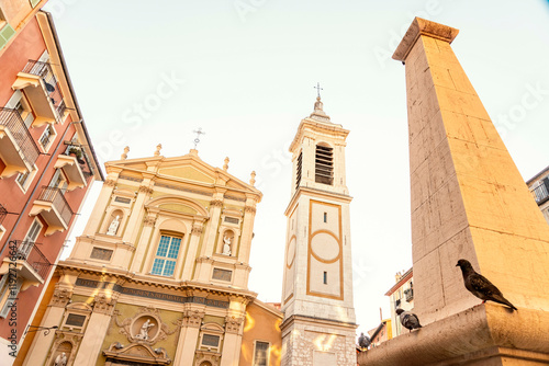 A pigeon perches on a sunlit ledge with a baroque church and an obelisk in the background against a clear blue sky, Nice, France photo