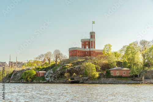 Red brick Stockholm Castle with a flag tower on an island against a clear blue sky with surrounding greenery, Stockholm, Sweden photo