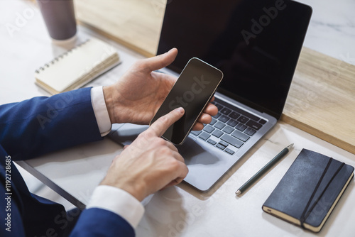 Fotografie Man engaged with his modern smartphone, laptop slightly out of focus nearby agai