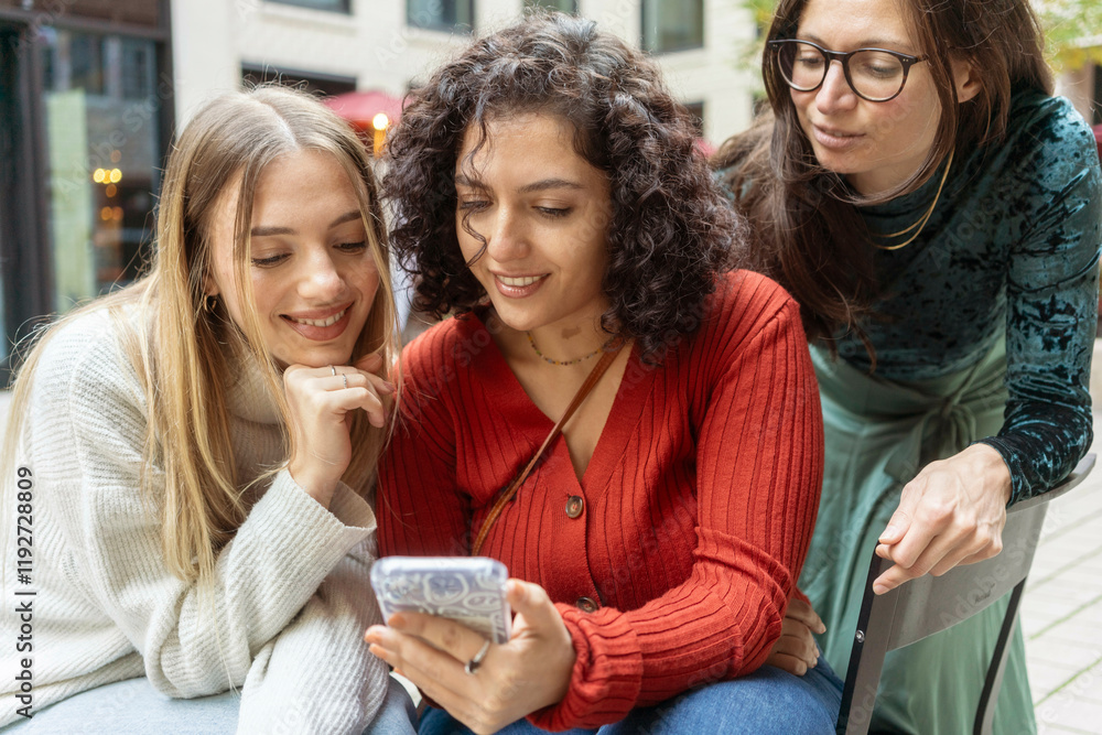 Three women share a moment looking at a smartphone with curiosity and amusement on a city street, Berlin, Germany Three women share a moment looking at a smartphone with curiosity and amusement on a city street, Berlin, Germany