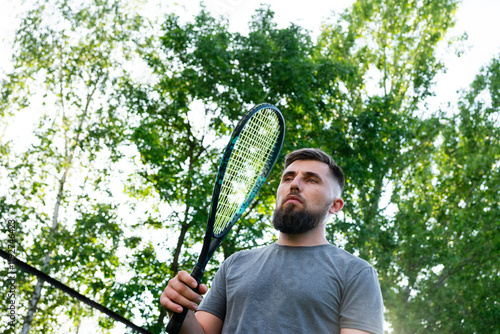 A man stands on a grassy area near a chain-link fence, holding a tennis racket and ball. He is dressed in sportswear and ready to play. photo