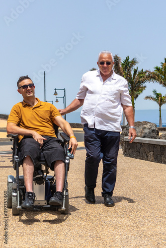 Senior man assisting adult male in wheelchair on sunny promenade photo