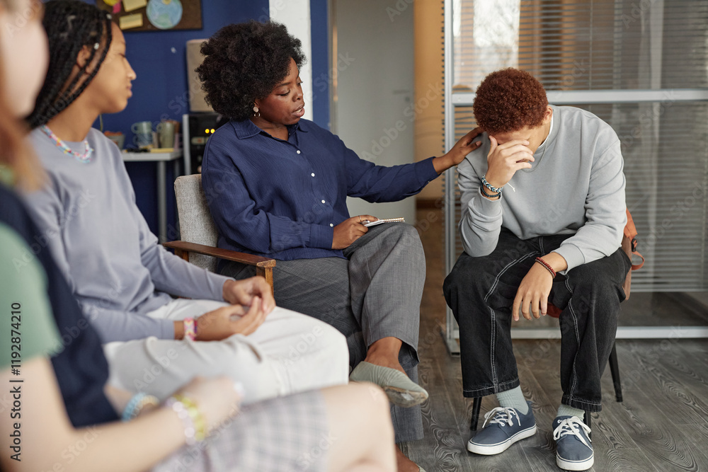 Full length portrait of teenage boy crying during support group meeting with African American woman therapist comforting him Full length portrait of teenage boy crying during support group meeting with African American woman therapist comforting him