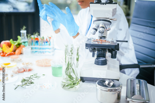 An African American female scientist works at her desk in a laboratory, closely examining a meat sample under a microscope.She focuses on modification,plant genetics,innovations in food meat research