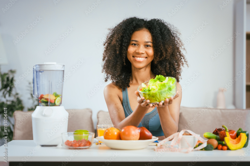 An African American woman in sportswear sits on a sofa near the dining table, preparing a healthy salad with vegetables fruits, drinking juice after exercise for weight control and low-fat nutrition An African American woman in sportswear sits on a sofa near the dining table, preparing a healthy salad with vegetables fruits, drinking juice after exercise for weight control and low-fat nutrition
