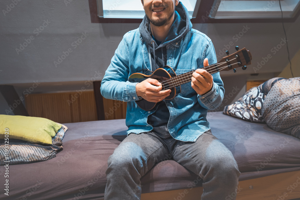 Obraz premium Musician with smile in a denim jacket and hoodie sits on a cozy bed, strumming a black ukulele. Enjoying the feeling of playing the ukulele and immersing oneself in the creative spirit