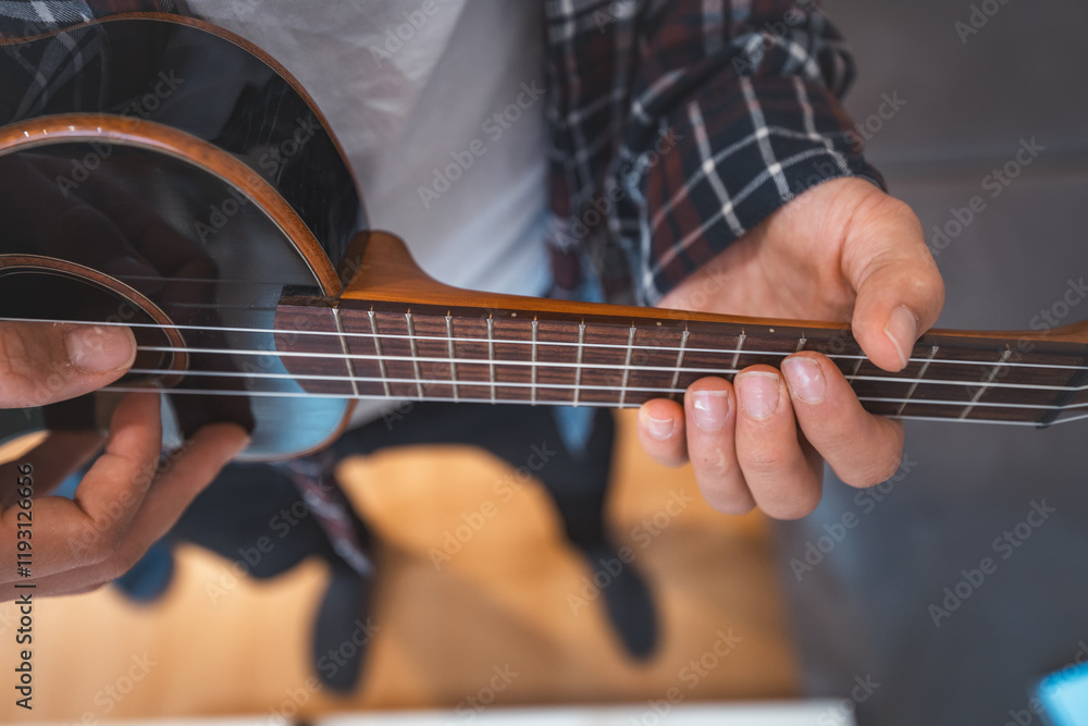 Fototapeta premium Close-up perspective captures a man's fingers plucking the strings of a glossy black ukulele. Making a music. Pleasure of playing an instrument