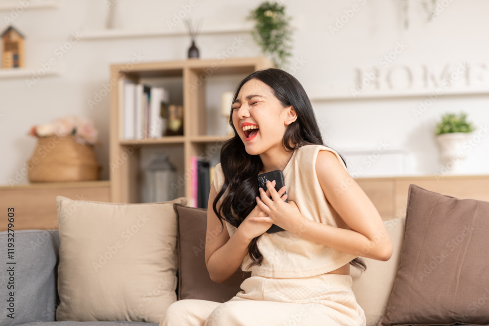 Woman laughing joyfully while holding smartphone on cozy sofa in bright living room Woman laughing joyfully while holding smartphone on cozy sofa in bright living room