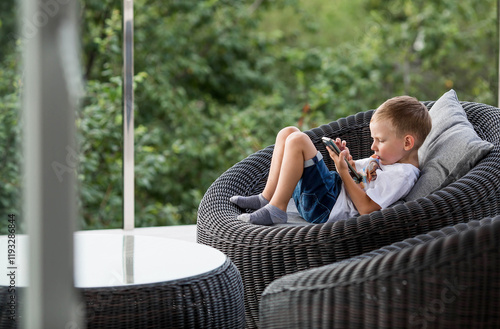 A 5-year-old little boy with a smartphone in his hands on the veranda. The child is playing on the phone. Harm of gadgets for children. Protect your child from the smartphone. Kids and gadgets.