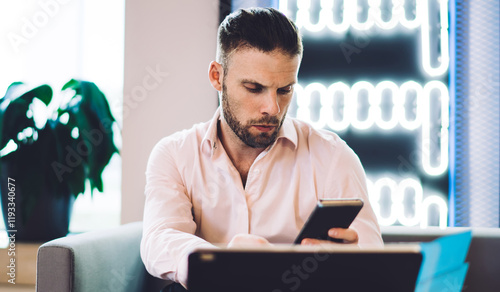 Caucasian man connecting to 4g wireless for reading received email message, serious male using cellphone device for online web messaging during remote job on portable touch pad in coworking interior photo