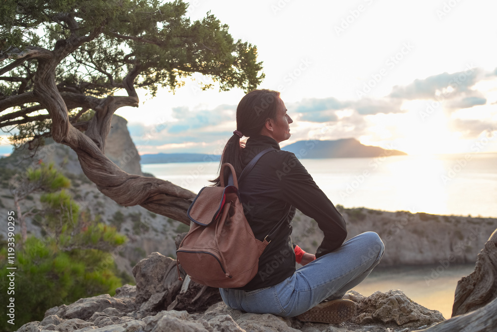 A young traveler with a backpack sits on the top of a mountain overlooking Cape Kapchik, Novy Svet village, Crimea, enjoying the view of the sea. A tourist with a backpack travels through the Crimea,
