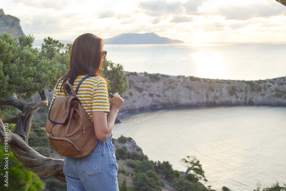 Happy female tourist with a backpack on the top of a mountain, enjoying the sea view, standing on a rock and looking into the distance. The village of Novy Svet in Crimea. Russia