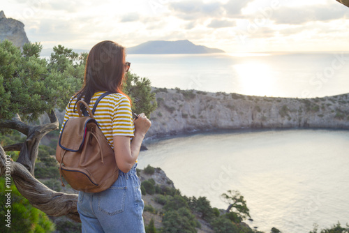 Happy female tourist with a backpack on the top of a mountain, enjoying the sea view, standing on a rock and looking into the distance. The village of Novy Svet in Crimea. Russia photo