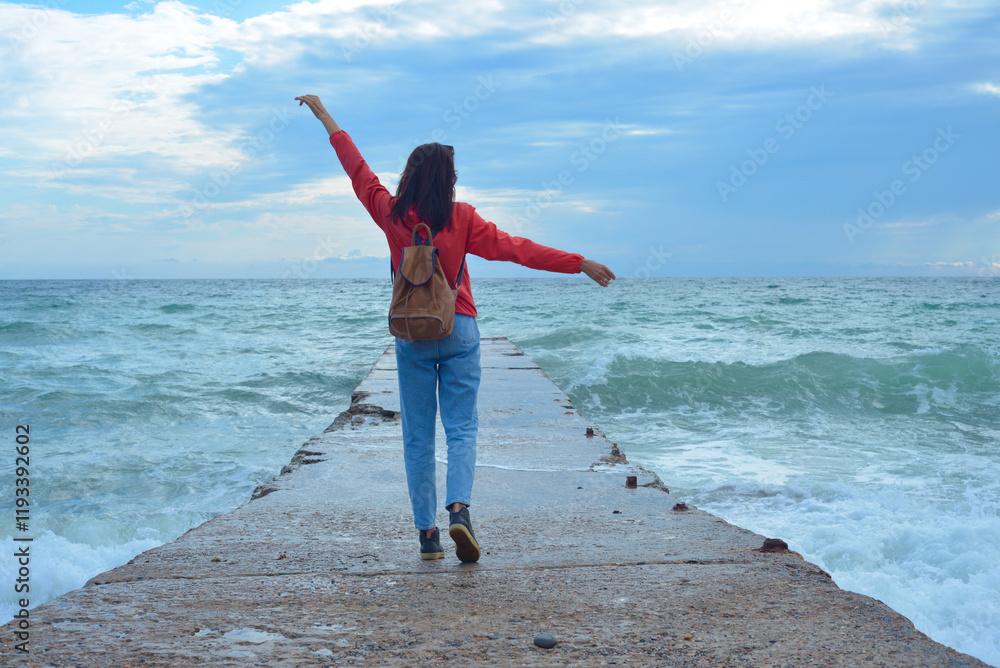Rear view of a young woman tourist with a backpack standing on the pier and waves hitting the pier in a storm. City of Sudak, Crimea.