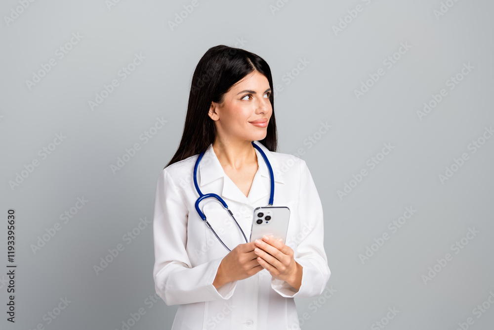 Young female doctor holding a smartphone in a professional white coat with stethoscope on a light grey background Young female doctor holding a smartphone in a professional white coat with stethoscope on a light grey background