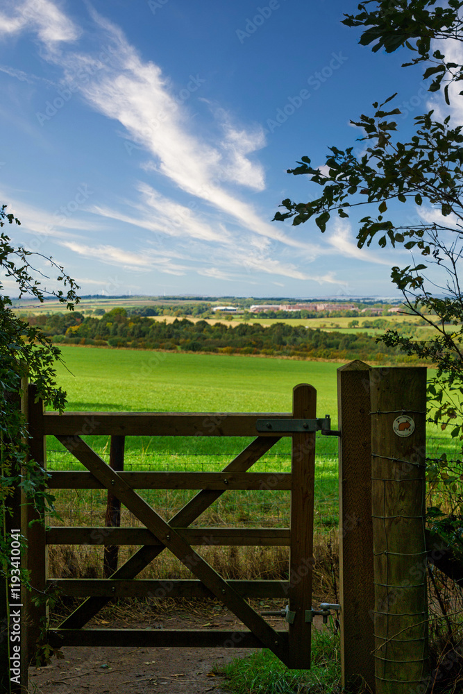 Fototapeta premium A rustic wooden gate opens to a picturesque landscape of rolling green fields and a bright blue sky, Harrogate, Yorkshire, UK.